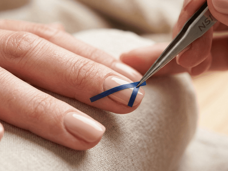 A close-up shot of tweezers placing thin striping tape in a geometric V-shape on a fingernail.
